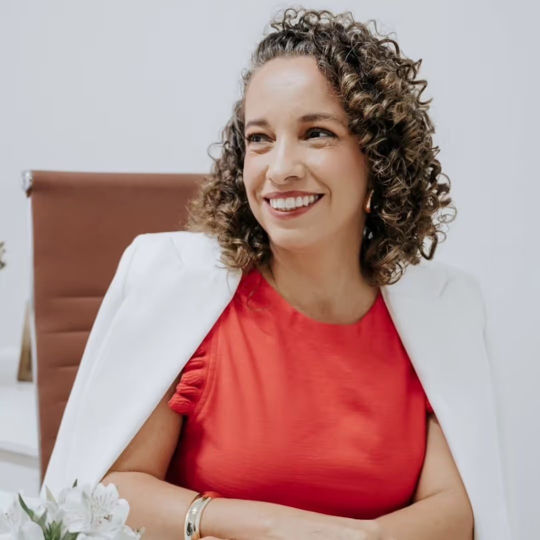 Heliane Pontes sorrindo, sentada em seu consultório com blazer branco